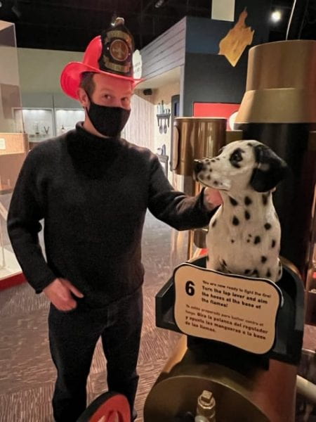 Josh trying his hand at a new career in the Chicago Fire Exhibit at the Chicago History Museum. Josh standing near Dalmation dog in the exhibit while wearing a Firefighter hat